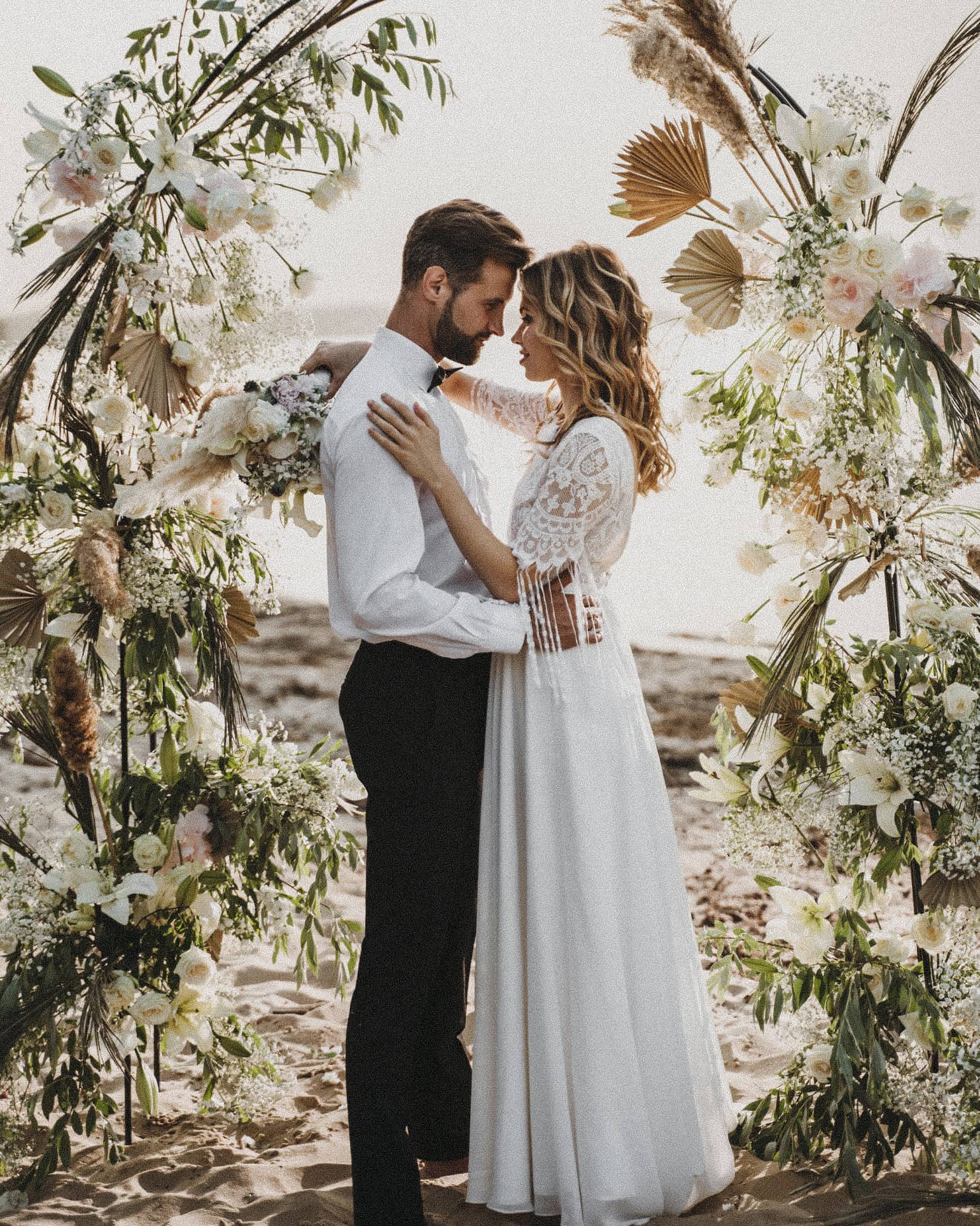 A couple embracing beneath an arch of dried grasses and white flowers on a sandy beach
