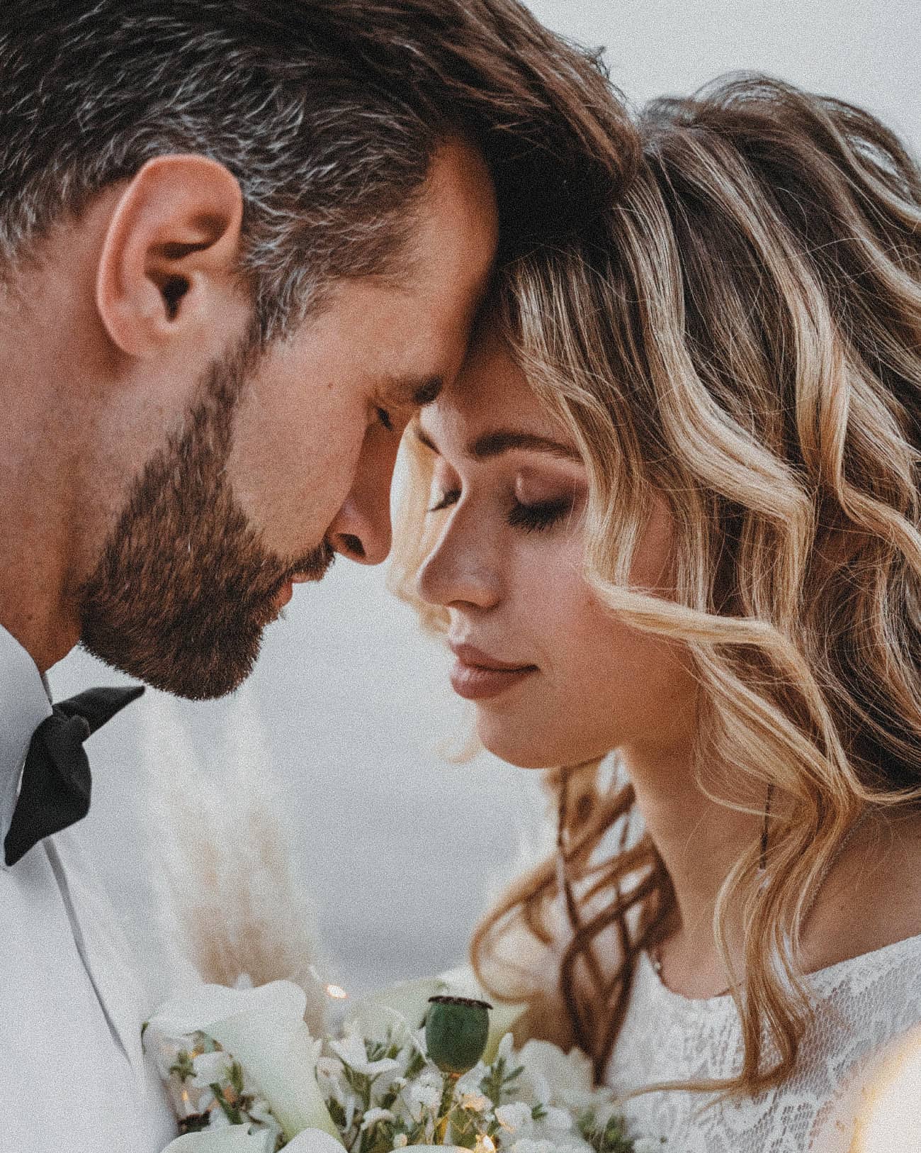 A bride and groom resting forehead to forehead, the bride with her eyes closed holding white flowers